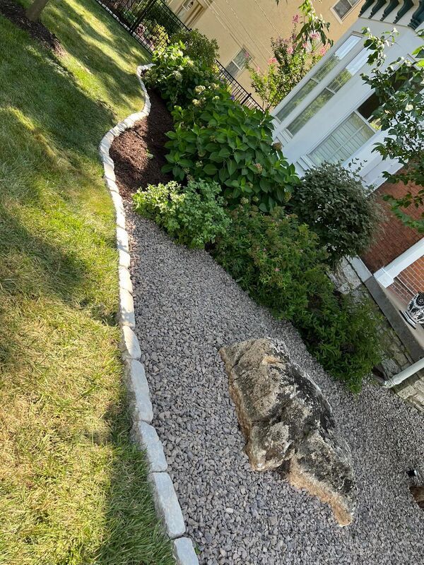 Gravel garden bed with a large rock, bordered by stones beside a white house and green lawn.