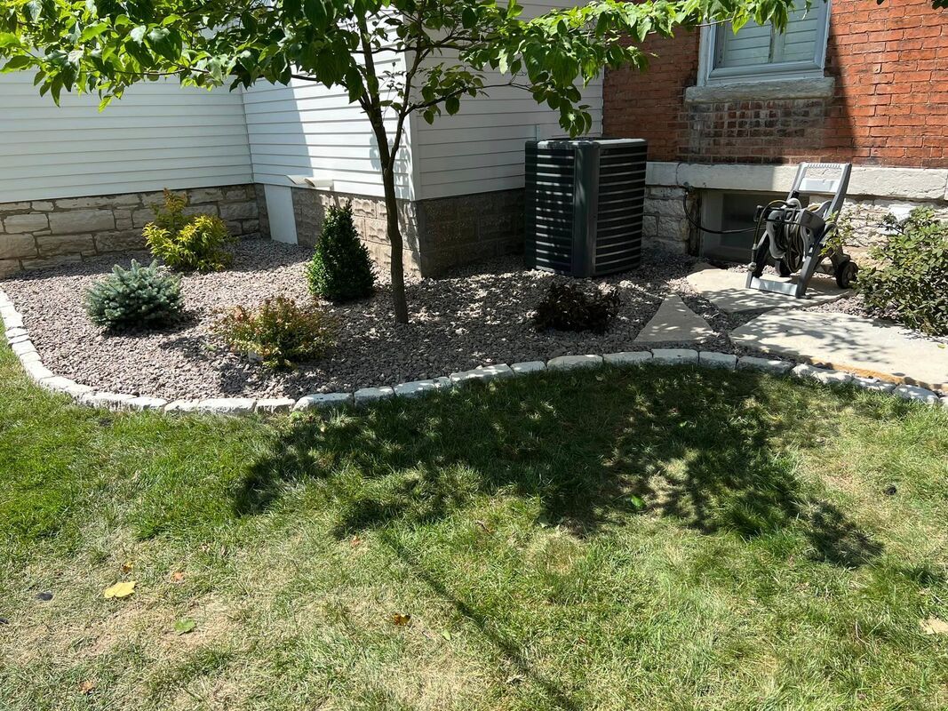 Front yard garden with shrubs, tree, white house siding, stone edging, and a bicycle near a brick wall