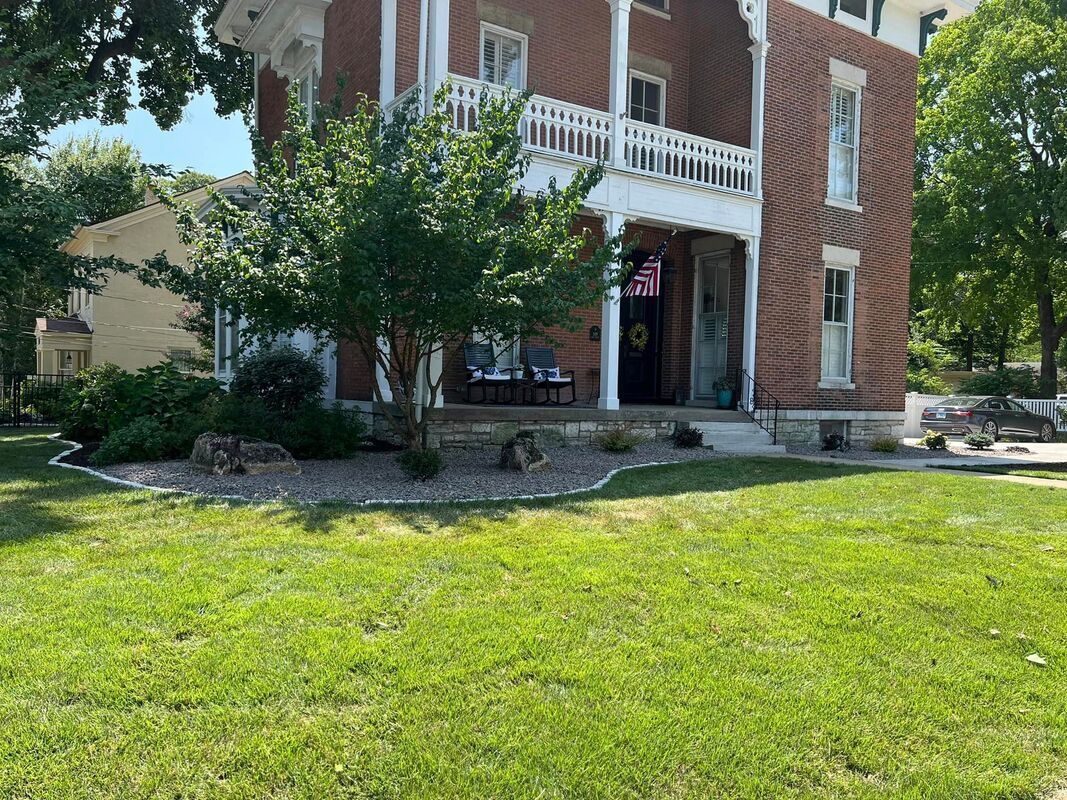 Brick house with white balcony and porch, set behind a green lawn and trees on a sunny day