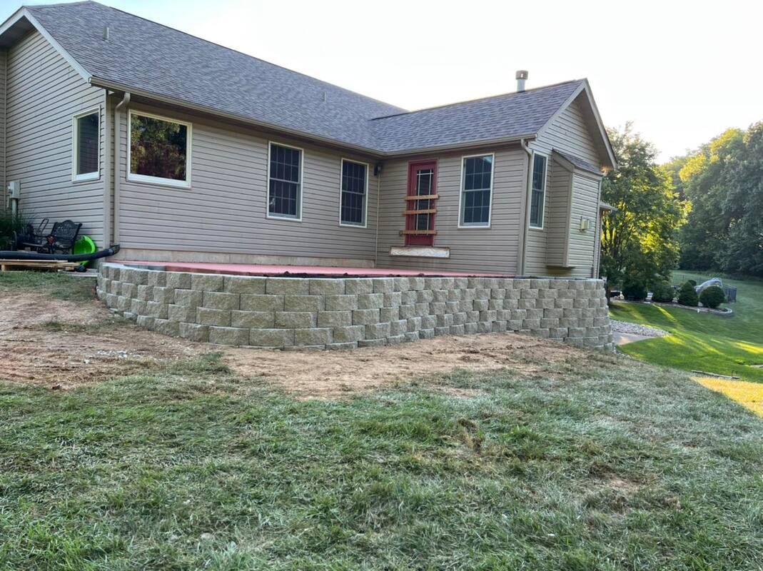 Side view of a beige house with a stone retaining wall and grassy yard at sunset