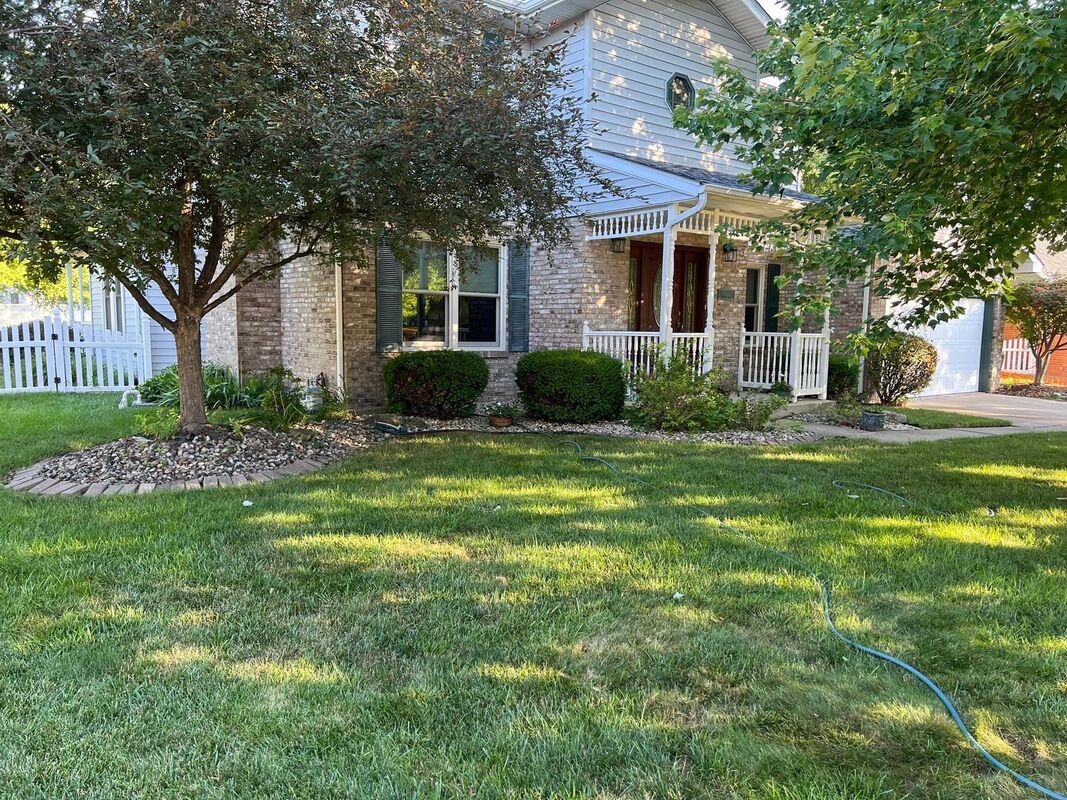 Brick house with a white porch, trees, and a green front lawn in sunlight