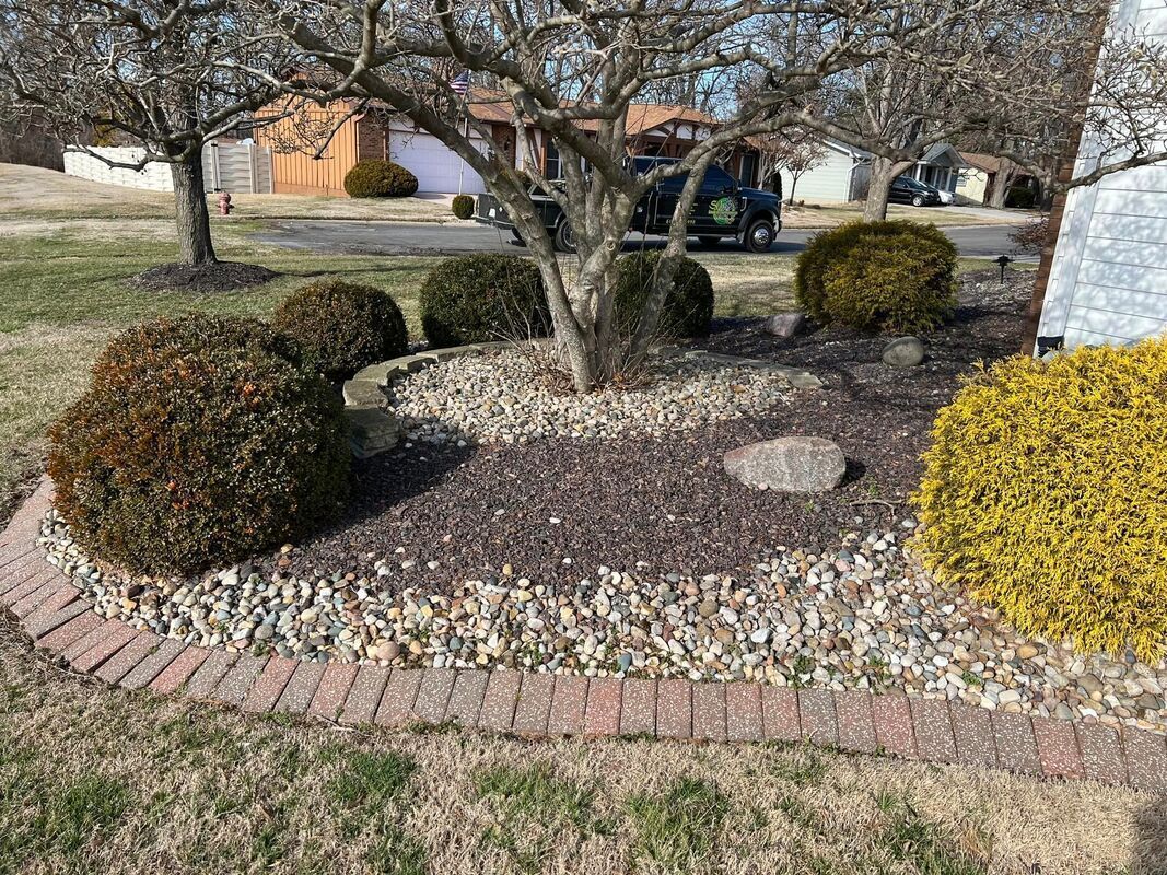 Landscaped front yard with mulch bed, trimmed shrubs, small tree, and brick border along a lawn.
