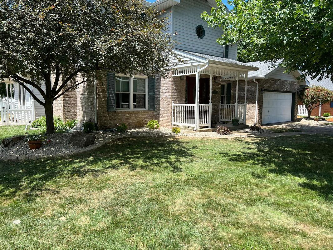 Suburban house with brick facade, white porch, garage, and a grassy front yard shaded by a tree