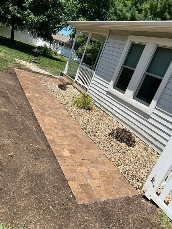 Brick walkway beside a house, bordered by mulch and gravel, with a lawn and trees in the background