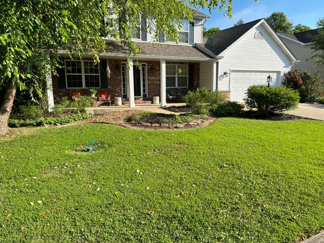 Suburban brick house with white garage, front porch, and a green lawn under trees