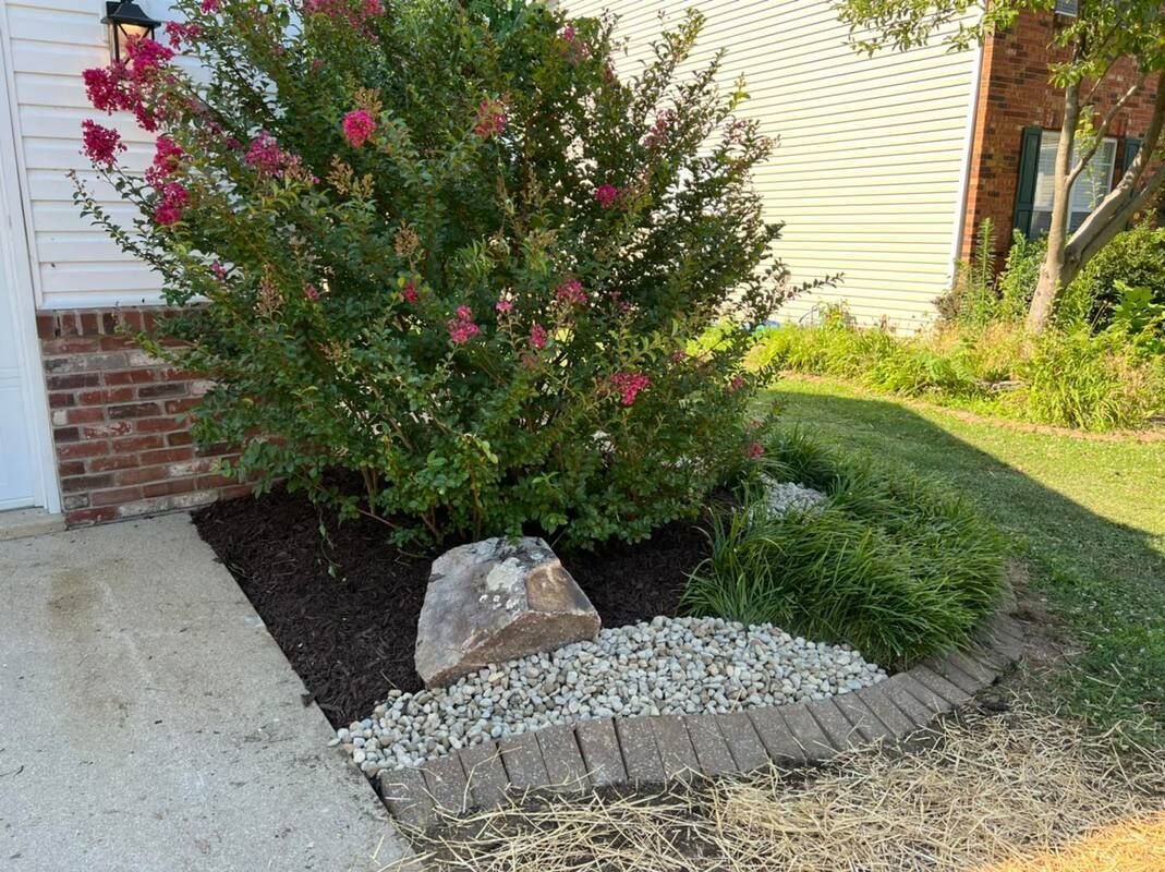 Front yard landscaping with flowering shrubs, rocks, and a curved brick border beside a house.