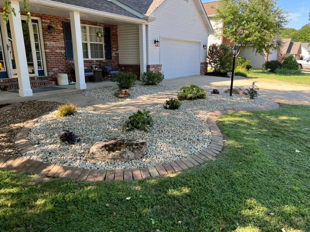 Front yard with stone garden beds, small shrubs, and a brick house with porch columns