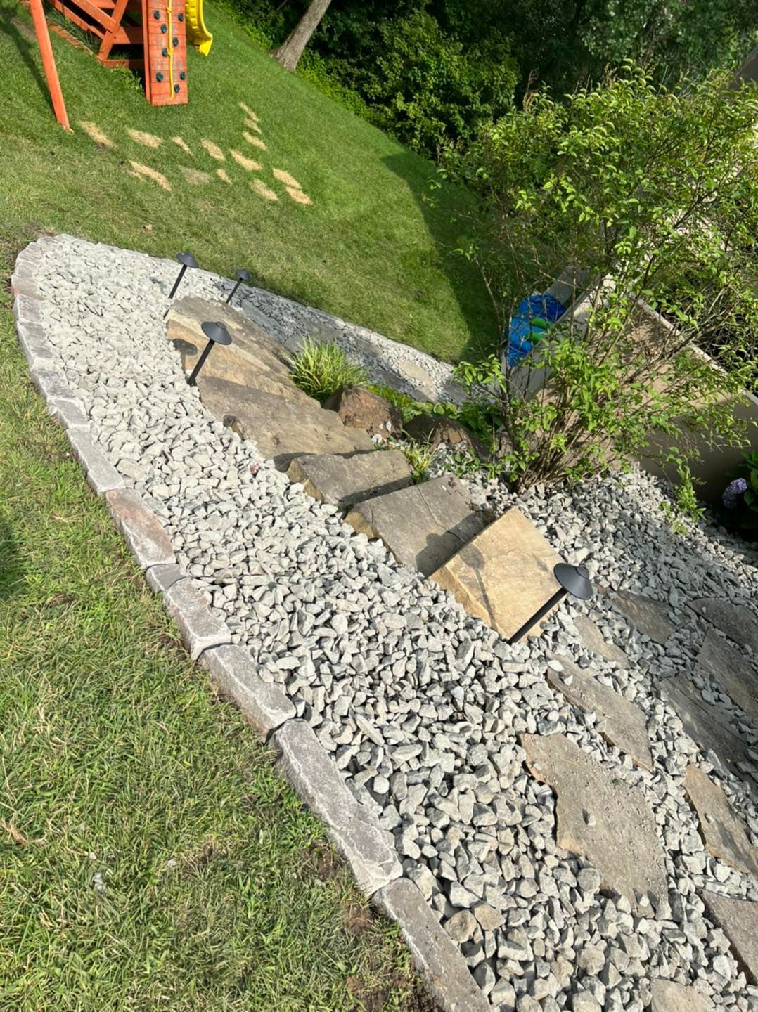Rock-lined garden bed with stepping stones, surrounded by grass and shrubs.