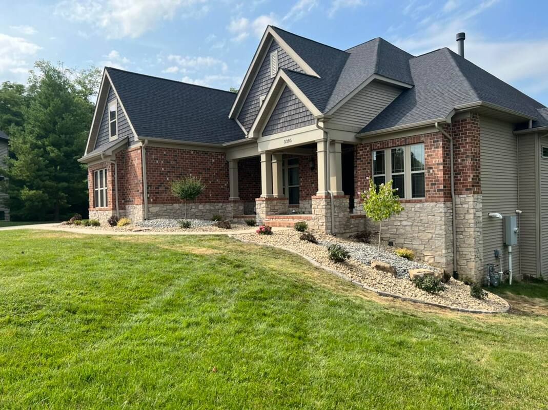 Suburban brick and stone house with dark roof and large green lawn under a blue sky