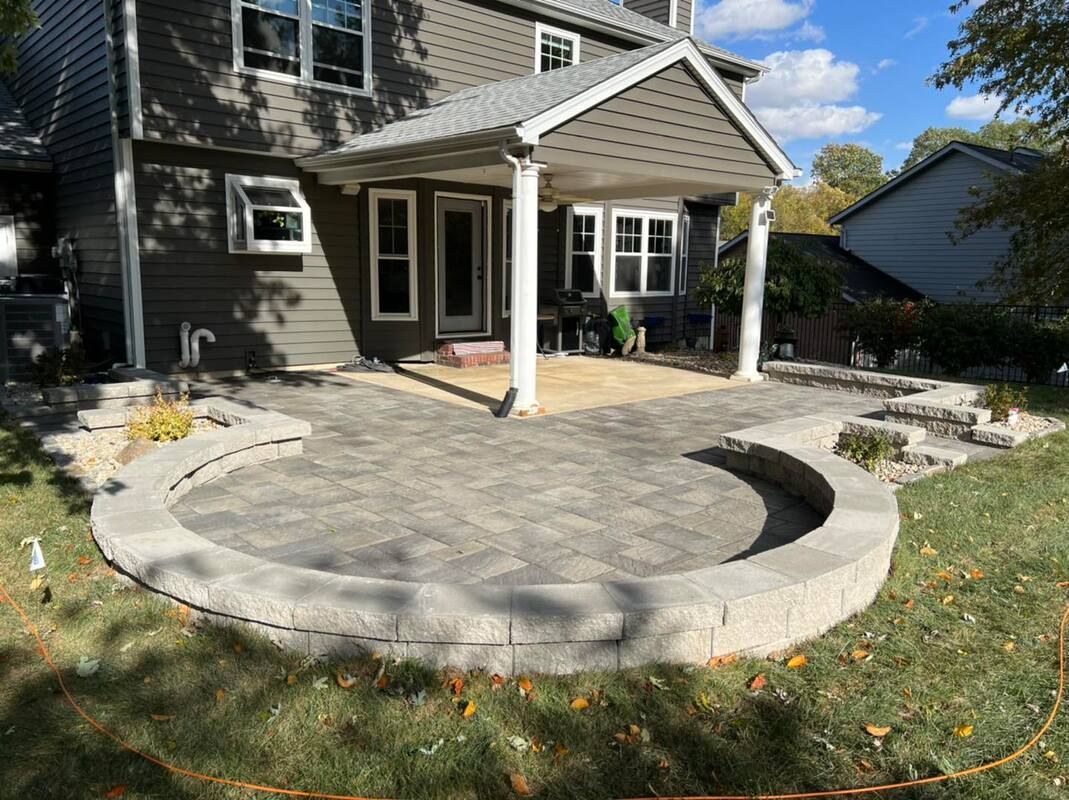 Front yard with a curved stone patio and covered porch beside a house, with landscaped rock beds and grass.