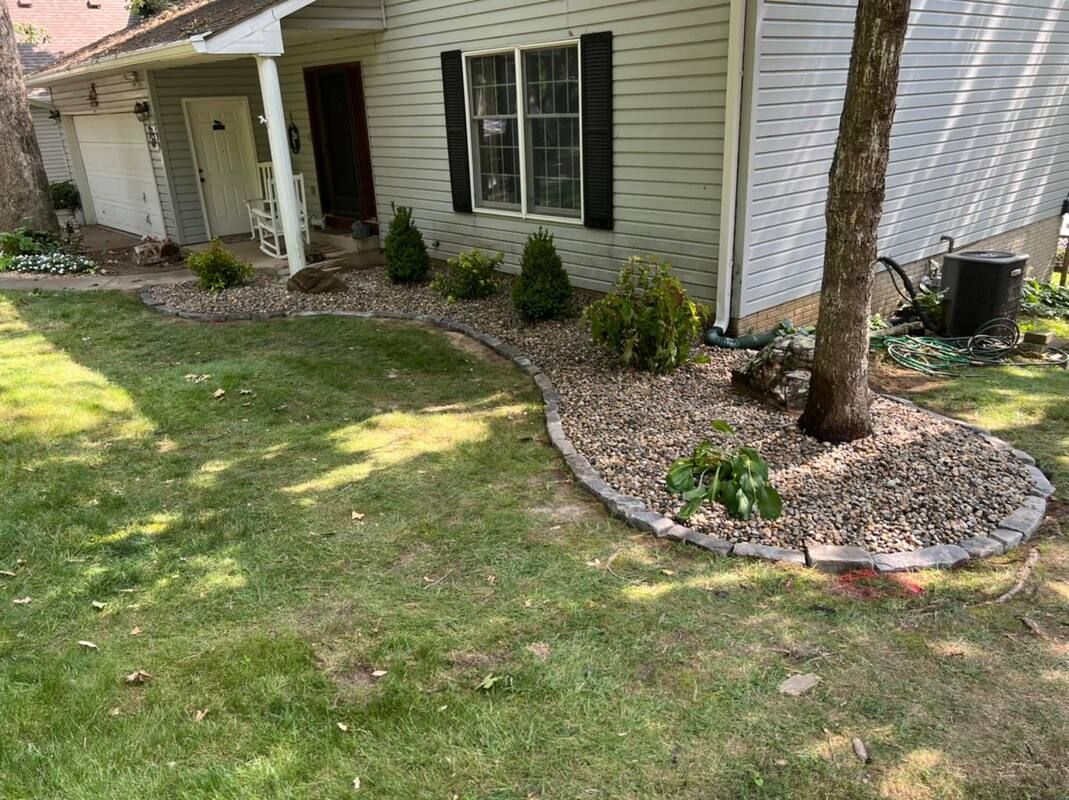 Front yard landscaping with a curved rock border, shrubs, and a tree beside a house.