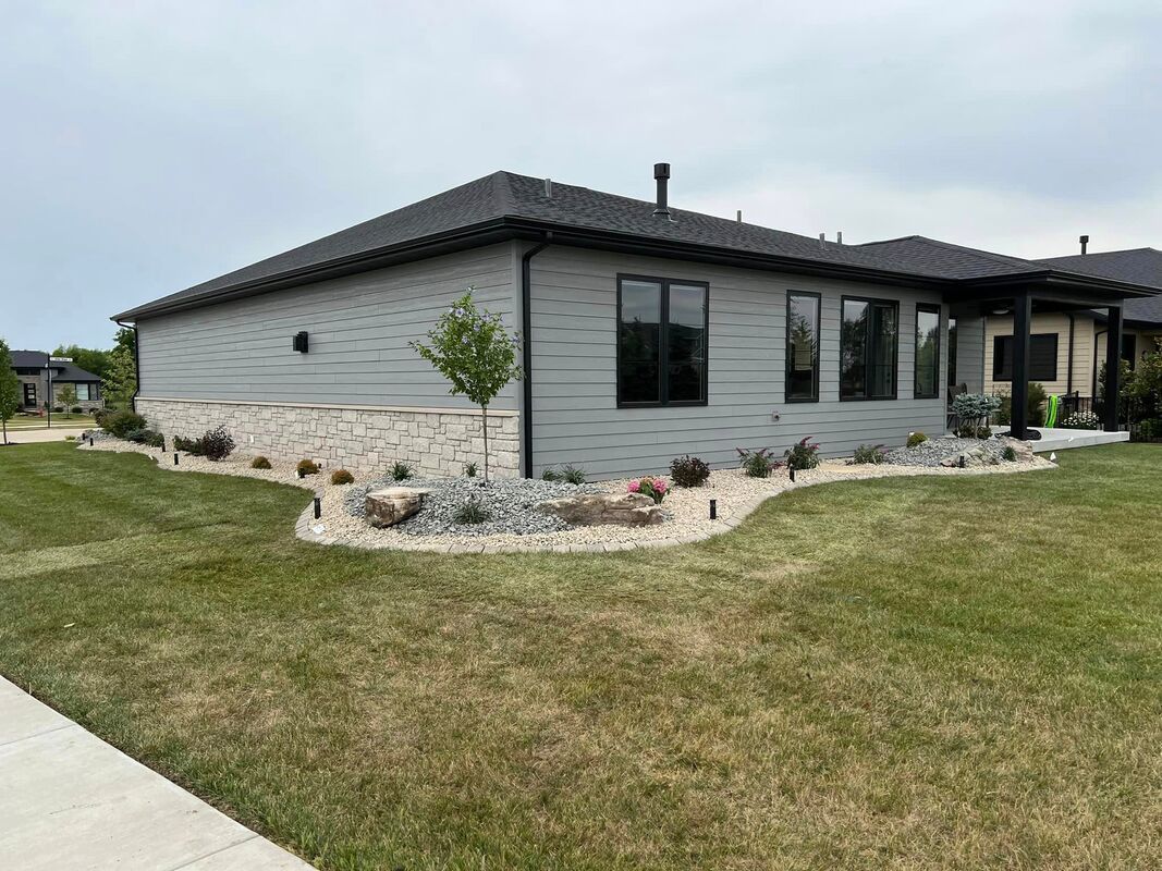 Single-story gray house with dark roof, stone foundation, and a landscaped lawn on a corner lot
