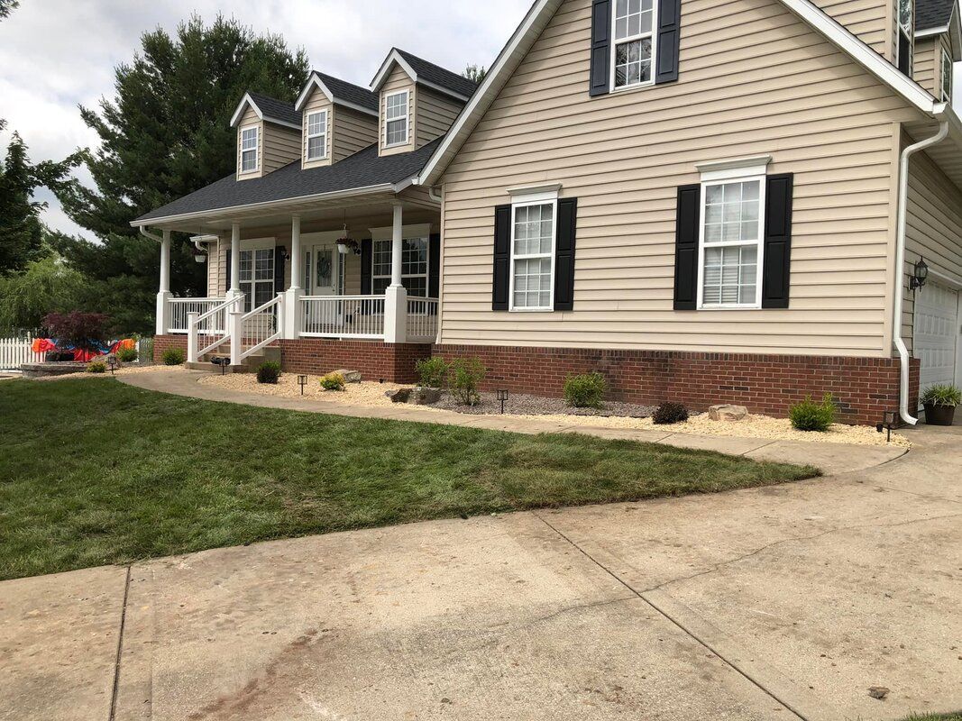 Suburban beige house with dark shutters, front porch, and driveway beside a green lawn