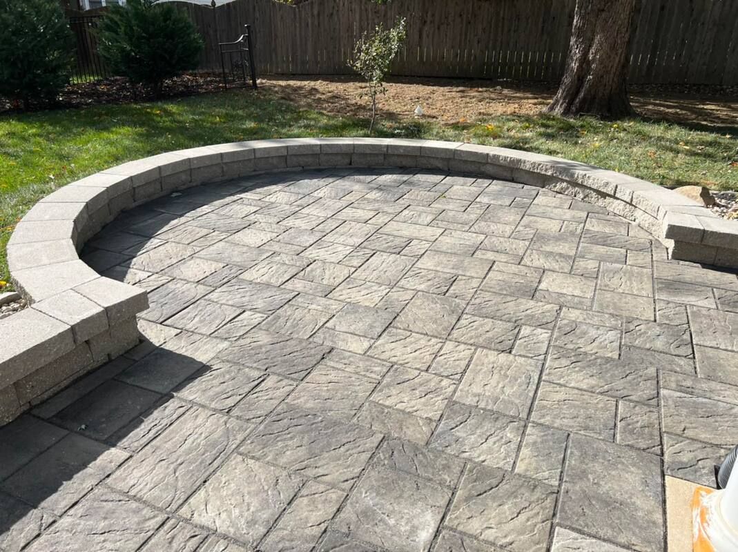 Curved stone patio with a low retaining wall and paver flooring in a backyard