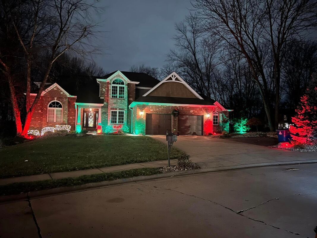 Suburban house at night decorated with red and green Christmas lights along the roof, windows, and yard trees
