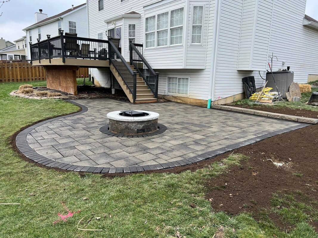 Backyard patio with stone pavers, circular fire pit, and stairs leading to a white house deck
