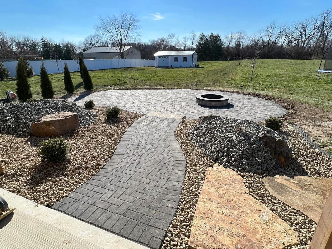 Curved stone path leading to a gravel fire pit area in a sunny backyard with lawn and a white shed