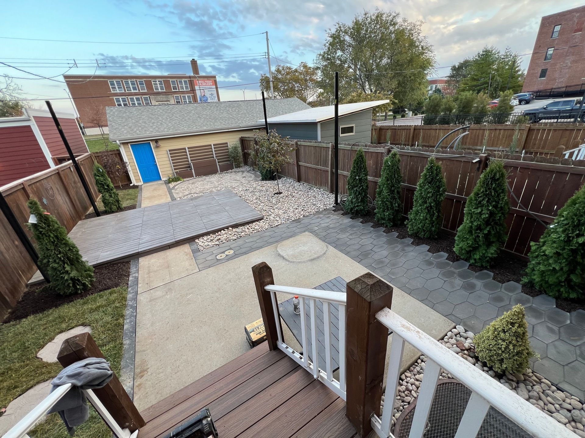 Backyard patio with wooden deck steps, stone pavers, fence, and rows of shrubs beside a shed