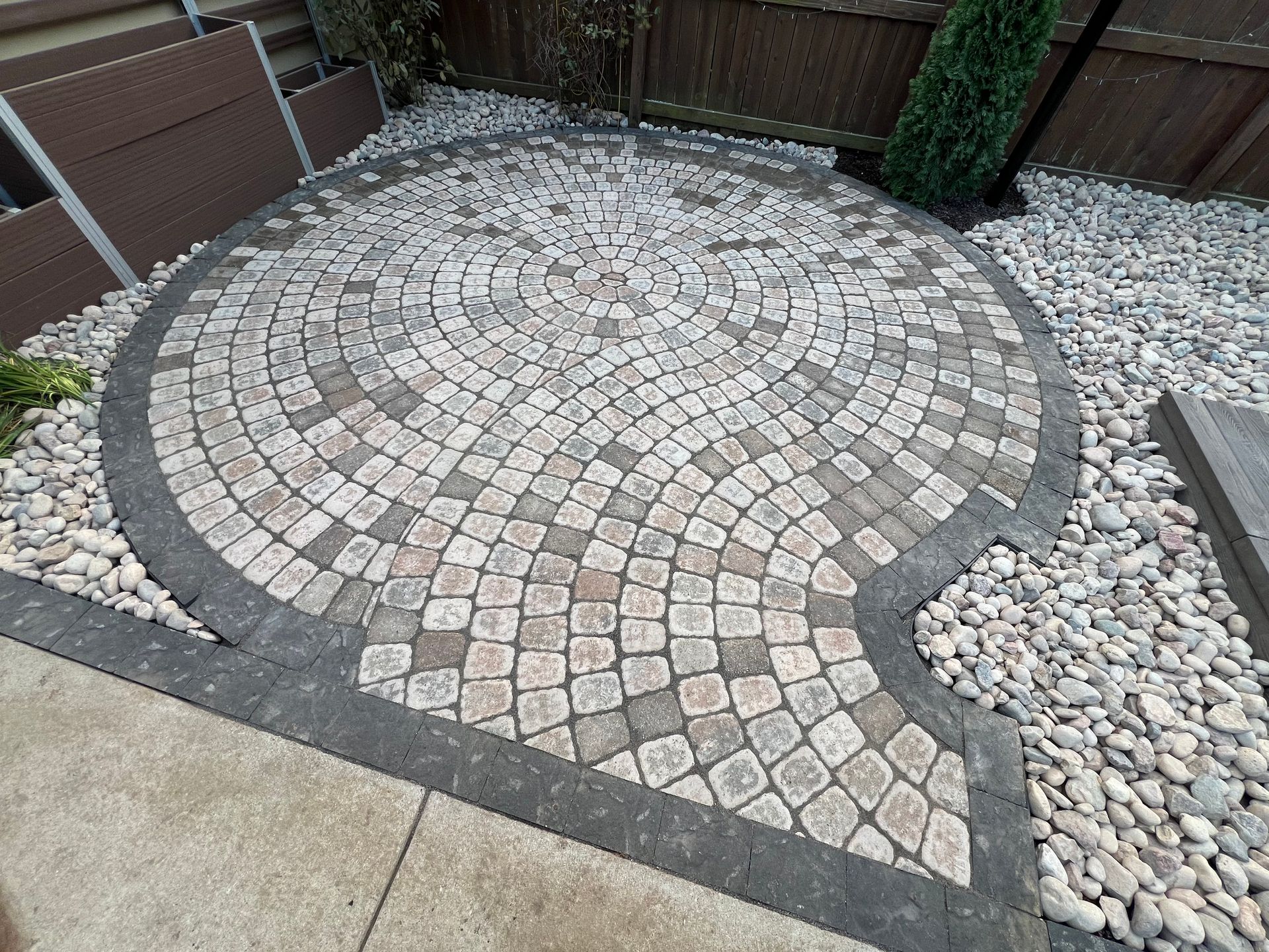 Circular brick patio with dark border stones in a gravel backyard beside a wooden fence