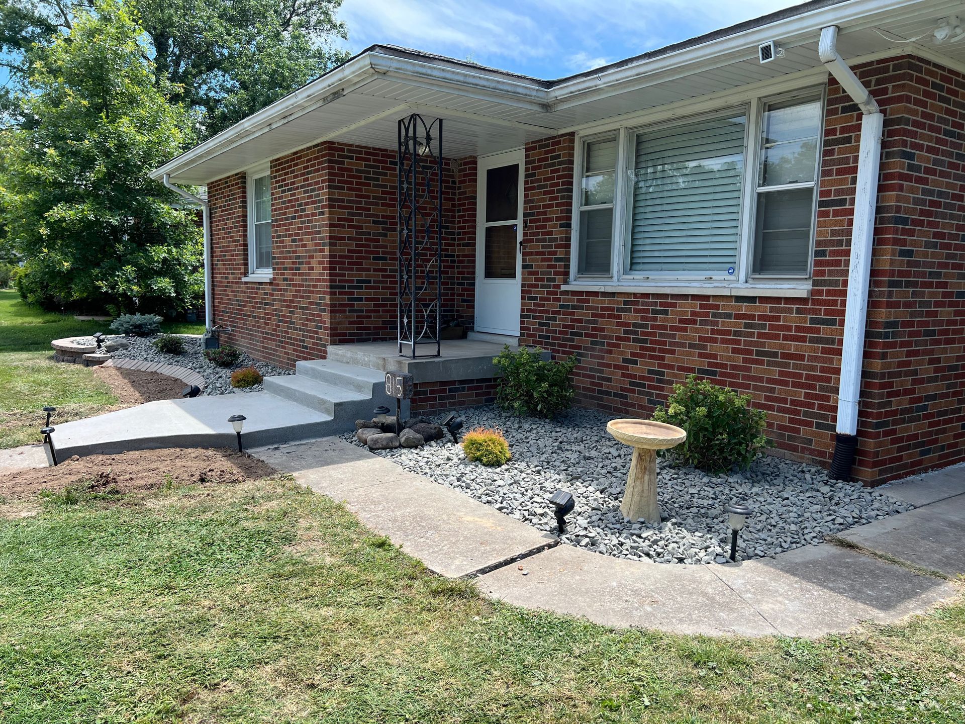 Brick house front entrance with concrete walkway, white steps, and small rock garden.