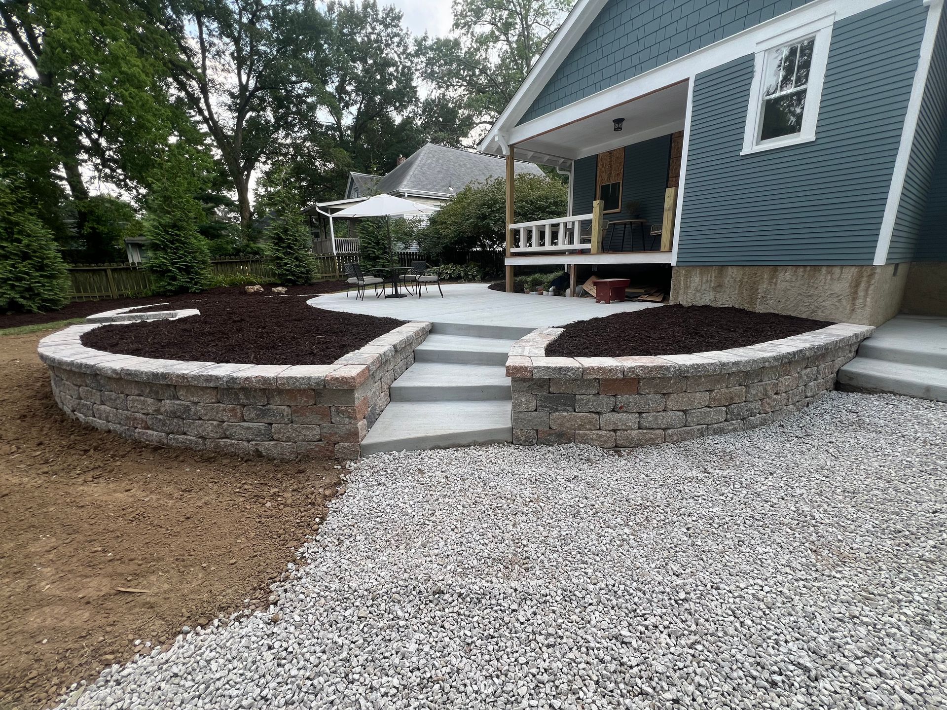 Curved stone retaining walls and steps lead to a blue house with a small porch and gravel path