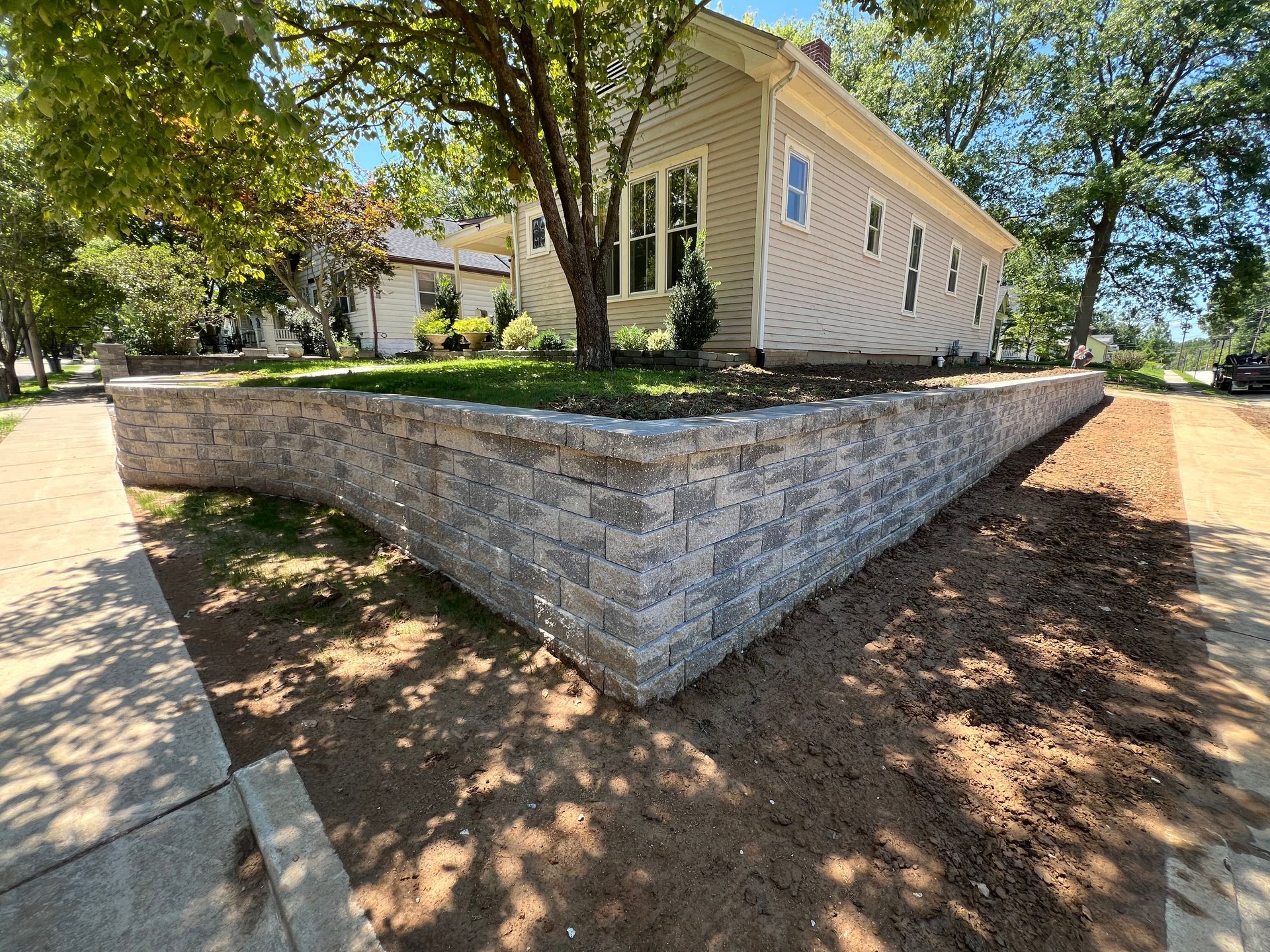 Single-story beige house with white shutters on a stone retaining wall under trees