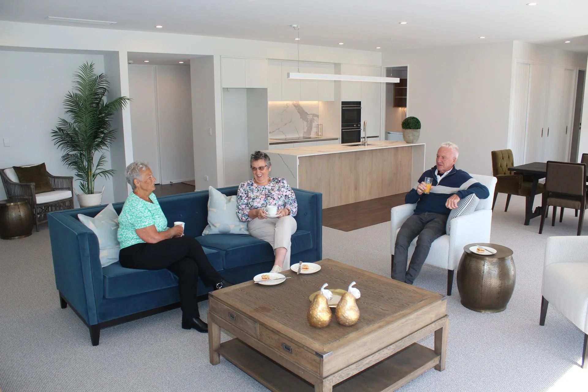 Three people sitting in a modern living room with a kitchen in the background; they are drinking beverages and conversing.