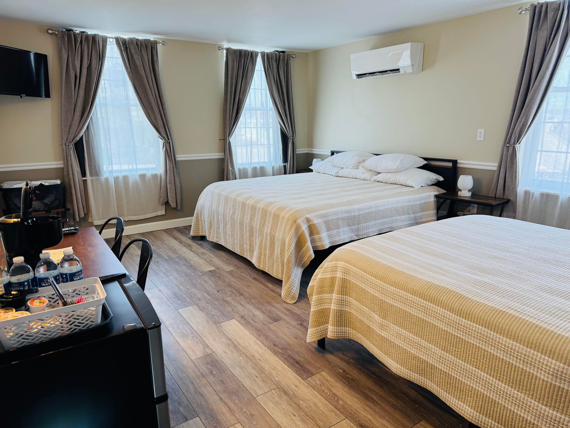 A hotel room with two beds, beige walls, wood floors, floor-length curtains, and a desk in the foreground.
