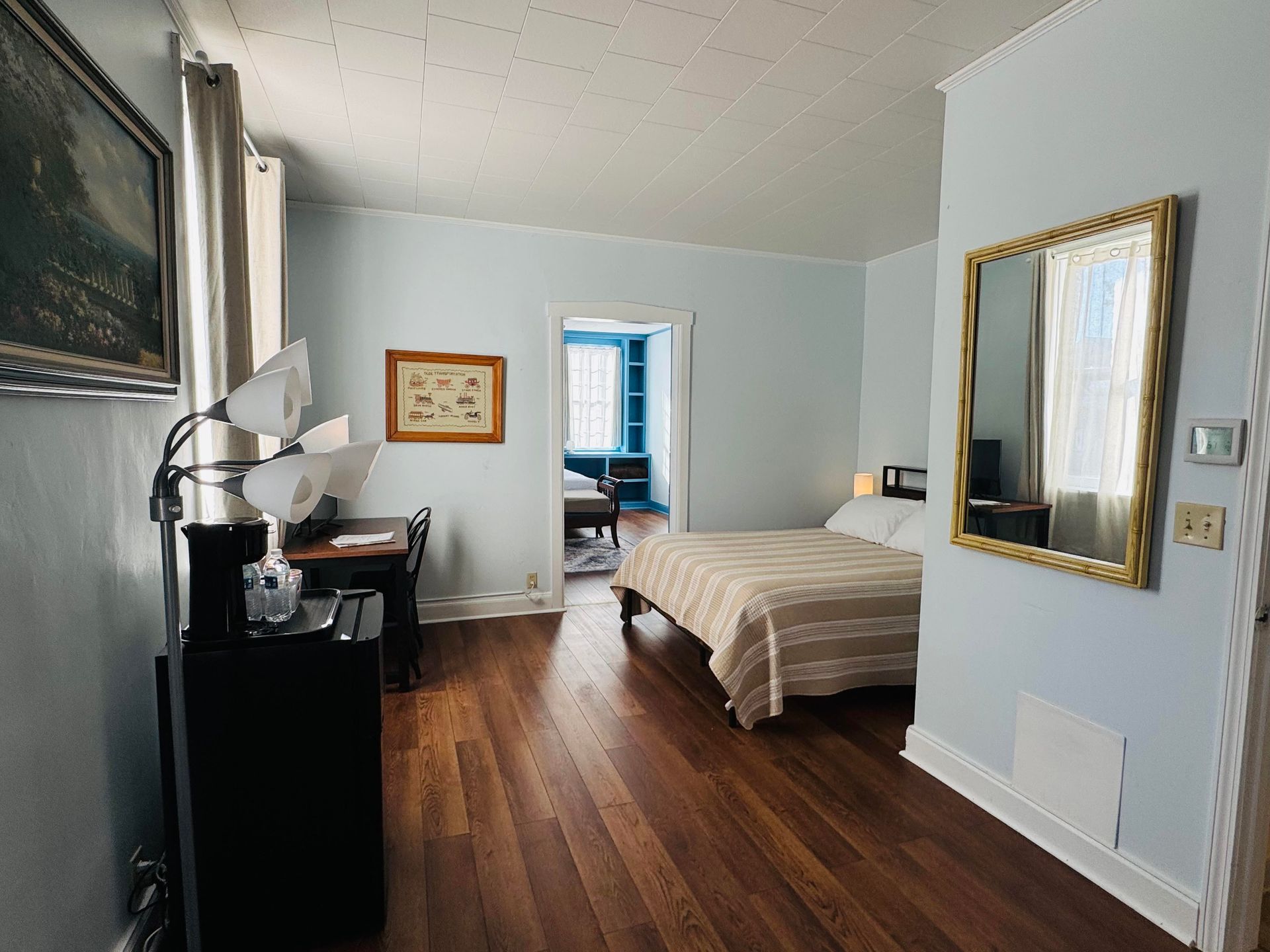 A bright bedroom with hardwood floors, a made bed, a desk in the corner, and a gold-framed mirror on a light blue wall.
