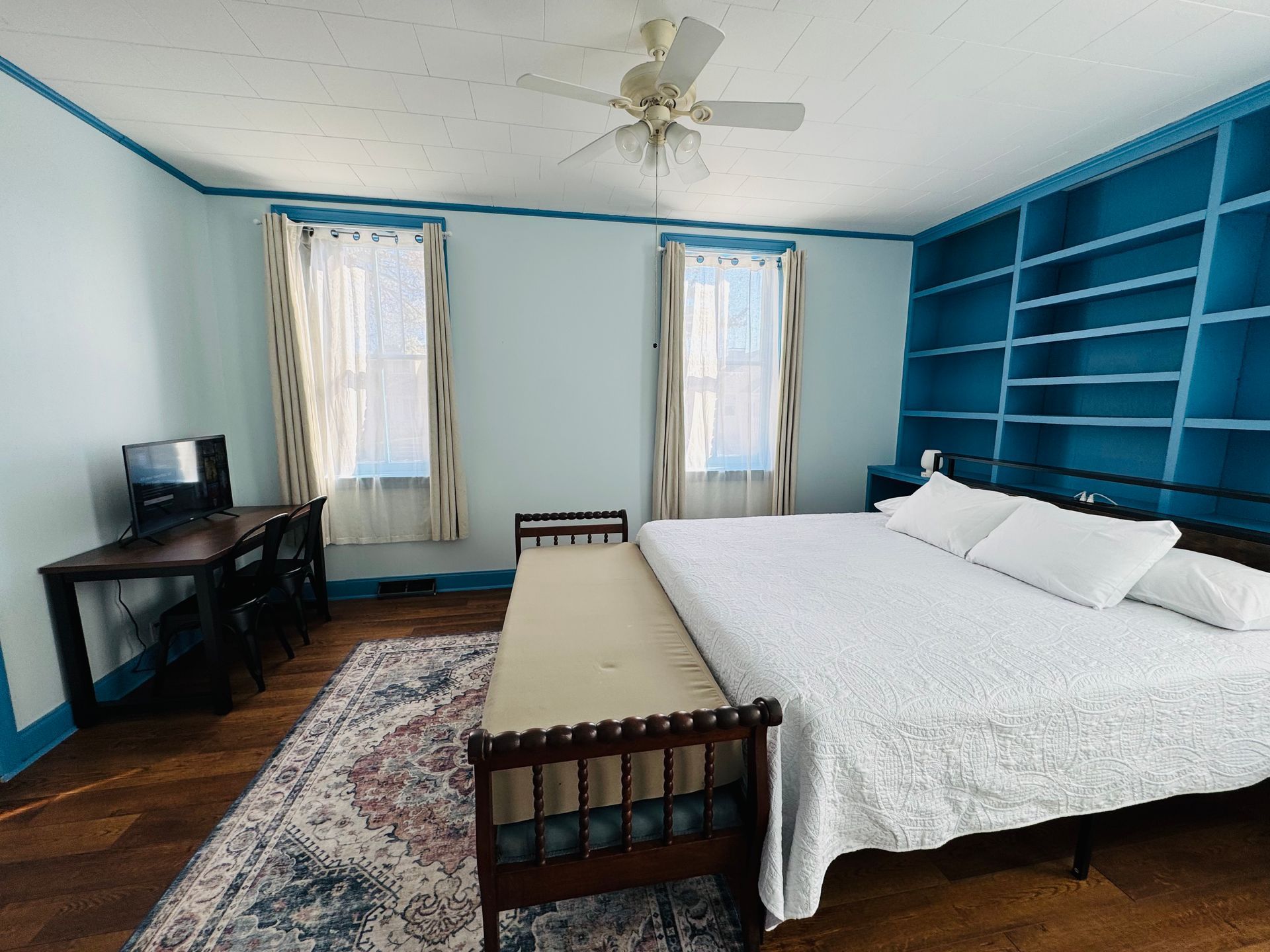 A bedroom with light blue walls, a large bed with white bedding, blue built-in shelves, a desk, and patterned area rug.