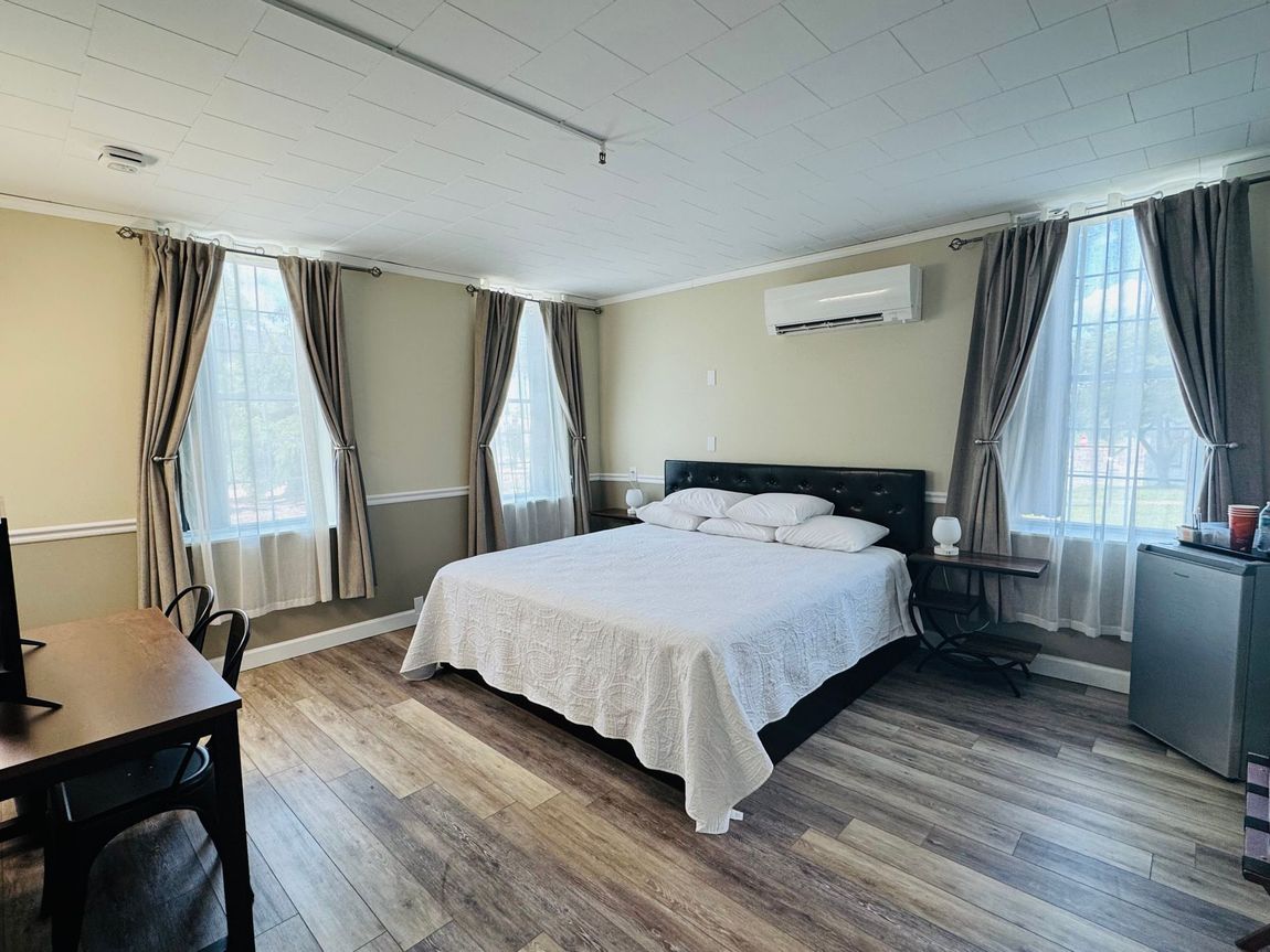 A bright, neutral-toned hotel room featuring a queen bed, wooden floors, two windows with curtains, and a desk.