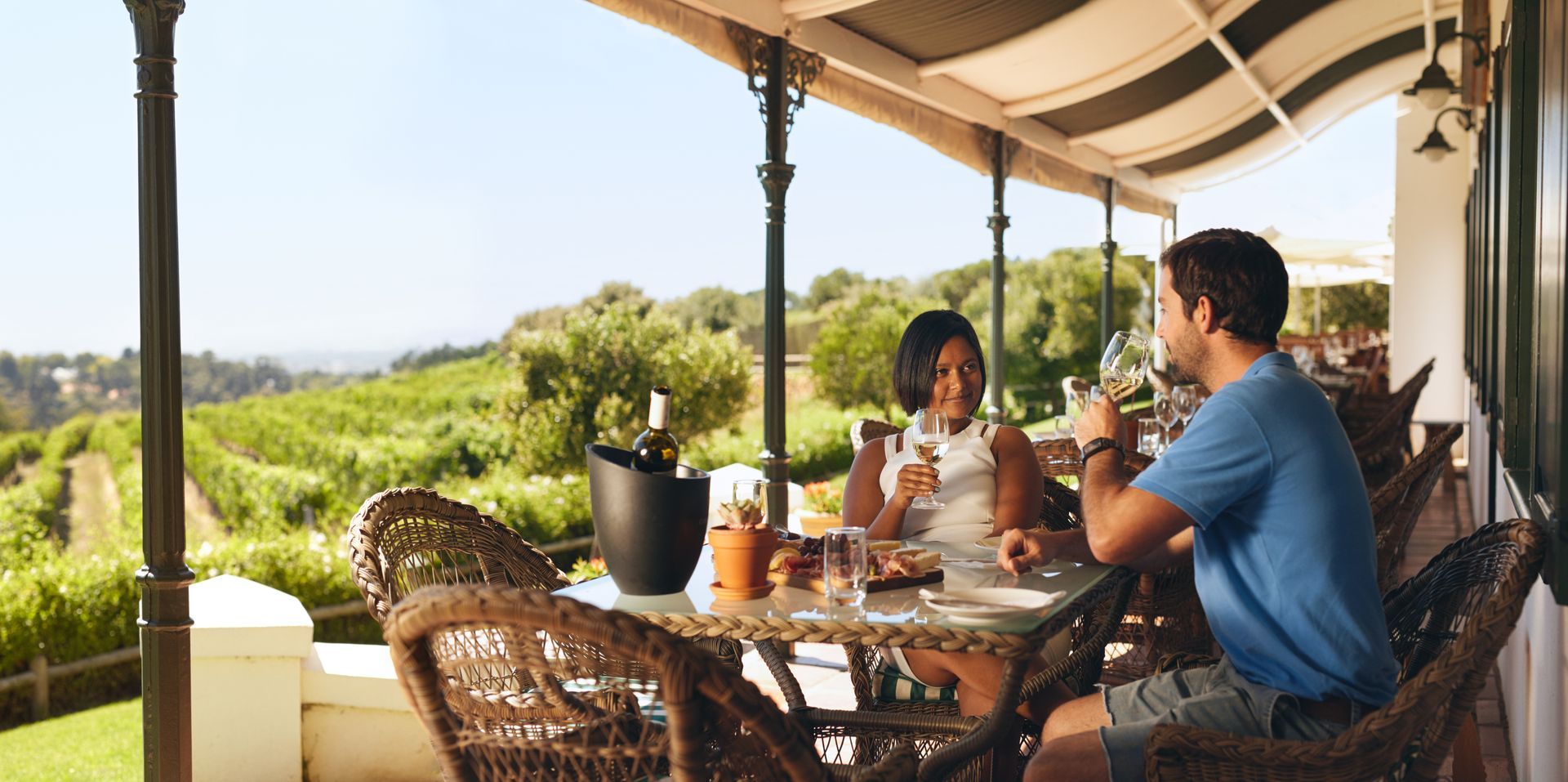Two people chatting at a wicker table on a covered patio overlooking a lush landscape.