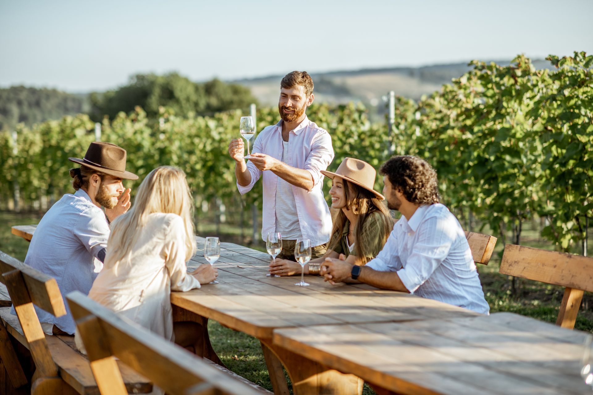 Friends chatting at a vineyard picnic table, with one person pouring wine outdoors.