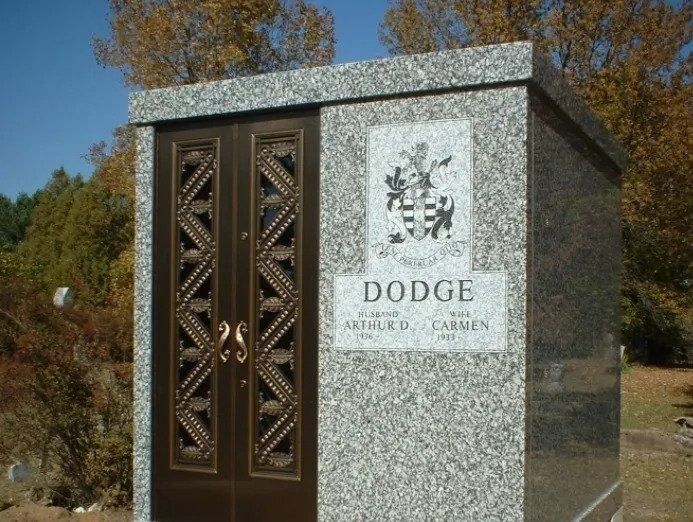 Granite memorial with bronze doors and a “DODGE” plaque outdoors among trees