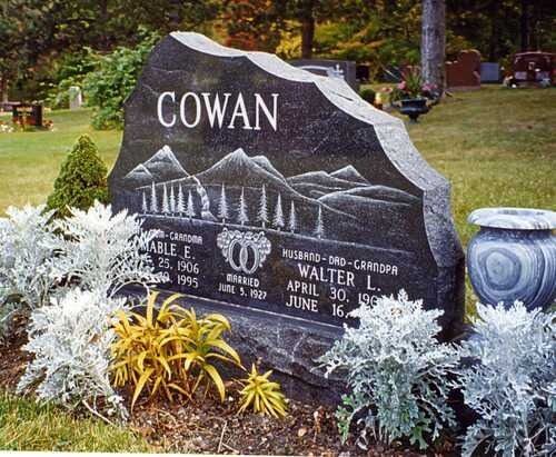 Black gravestone with “COWAN,” mountain engraving, and white ornamental plants in a cemetery