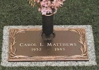 Gravestone for Carol L. Matthews, 1932–1985, with pink flowers above the marker