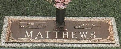 Brown granite grave marker engraved “MATTHEWS” with two name plaques and flowers in a vase on top