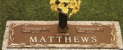 Brown grave marker with the name “Matthews” and small yellow flowers above it