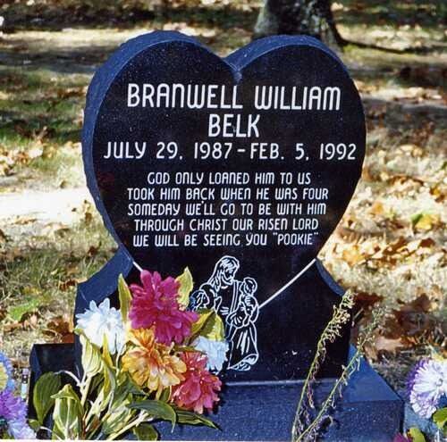 Gravestone with engraved text and flowers in front, set outdoors among grass and trees