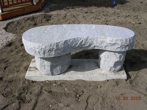 Rough white stone bench with curved top and two carved supports on a concrete base outdoors on dirt
