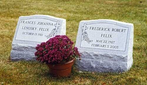 Two engraved cemetery headstones with a purple flower pot in front on a grassy lawn.