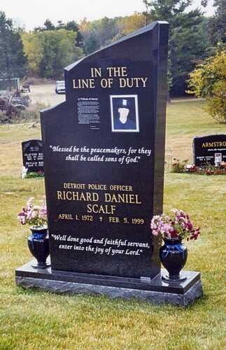 Black memorial grave marker with flowers in a cemetery, reading “In the Line of Duty”