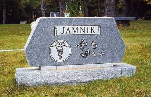Stone memorial marker reading “JAMNIK” in a grassy cemetery, with a crucifix and butterfly engravings.