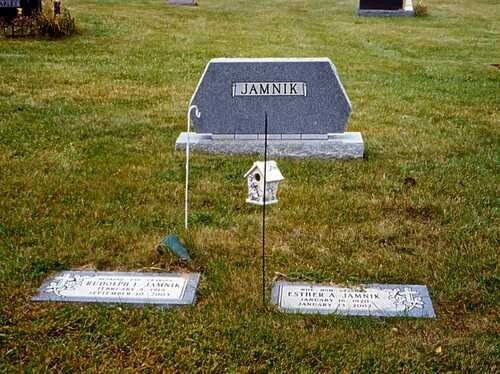 Gravestone in a grassy cemetery plot with small memorial markers and a white lantern in front