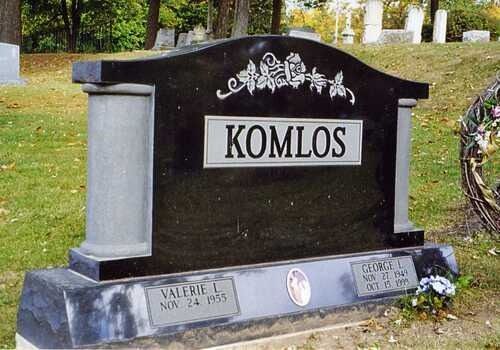 Black gravestone with “KOMLOS” in a cemetery, with decorative floral engraving and plaques at the base