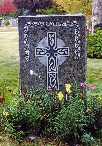 Gravestone with ornate Celtic cross engraving, surrounded by flowers in a cemetery garden.
