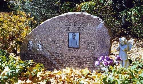 Memorial plaque in a flower-filled garden with a small portrait and angel statue beside it