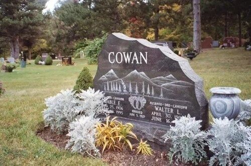 Gravestone marked “COWAN” in a landscaped cemetery, surrounded by flowers and greenery.