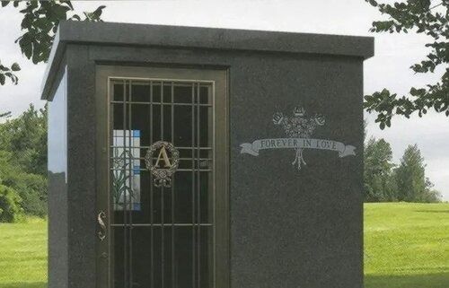 Small dark stone mausoleum with barred door and emblem, set in a grassy cemetery.