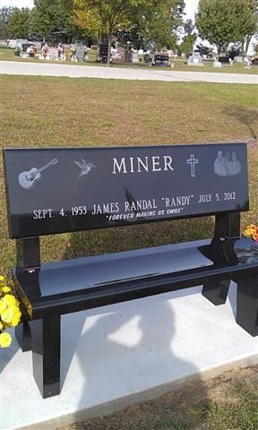 Black memorial bench with silver plaque and guitar decoration in a park.
